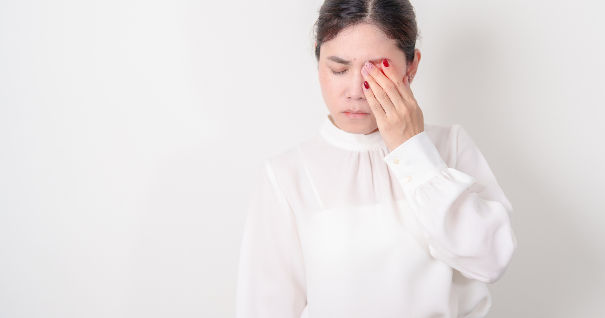 Fatigued woman with diabetes rubs eyes, standing against pale wall in white blouse, depicting late afternoon energy crash for diabetes fatigue article