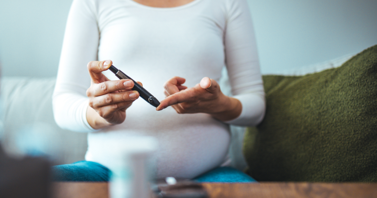 Pregnant woman seated and using a lancing device on her fingertip to check blood sugar levels at home.
